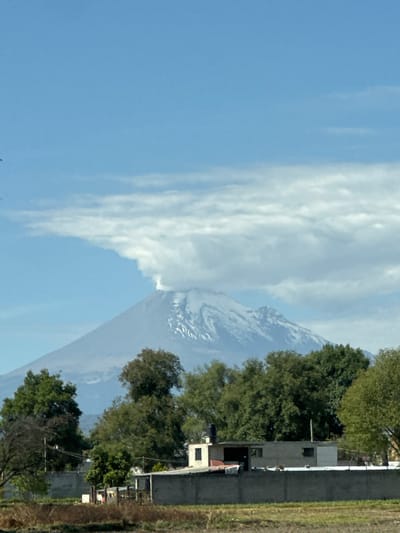 A landscape in San Rafael Tlanalapan, Mexico, shows a snow-capped volcano rising into a blue sky with scattered white clouds. The right side of the image reveals green trees and a two-story grey building adjacent to a field with dry vegetation. No people are visible.