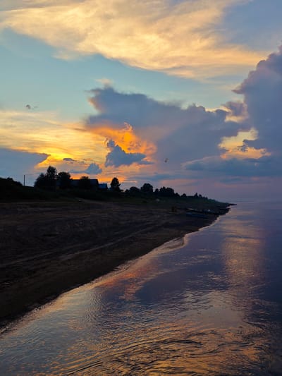This media file captures a serene sunset over a tranquil body of water, likely a river or lake, with a sandy shoreline in the foreground. The sky is a canvas of vibrant colors, with streaks of orange, yellow, and pink clouds set against a soft blue backdrop. Large, dramatic clouds, some tinged with gray and purple, add depth and texture to the scene.
On the shoreline, a dark, sandy bank slopes gently towards the water. Tire tracks or other imprints are visible on the sand, suggesting past human or vehicle activity. Further inland, a line of trees forms a dark silhouette against the colorful sky, with several houses or buildings nestled amongst them. A few small boats are beached along the water's edge.
The water itself is calm, reflecting the warm hues of the sunset and the dramatic clouds above, creating a shimmering, abstract pattern on its surface. The overall mood is peaceful and contemplative, evoking the quiet beauty of nature at dusk. The time of day is clearly sunset, given the vibrant colors in the sky and the reflections on the water.
The location context provided places this scene in Vorogovo, Russia. There is no visible text in the image.