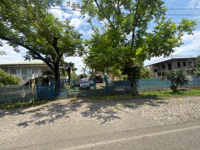 The image depicts a sunny day in სუფსის თემი, Georgia, with a road in the foreground and two houses behind a blue metal fence. The house on the left is a light-colored, two-story building with a green roof and visible windows and balconies. The house on the right appears to be under construction, with a stone facade and dark window openings. Several trees, including large deciduous trees and palm trees, are scattered throughout the scene, casting shadows on the gravelly ground. A blue gate provides access to the property. The sky is partly cloudy with white cumulus clouds. No people are visible in the image.