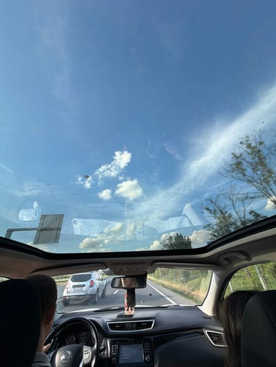 The image depicts a view from inside a car, likely the passenger seat, with the driver visible in the foreground. The car is on a road, and the rearview mirror shows another car, a silver SUV with the license plate "IF-53LWW", behind it. The dashboard of the car is visible, including the steering wheel and the infotainment system. Through the car's sunroof, the sky with scattered clouds is visible. Based on the visible road sign, the car is located in Frătești, Romania.