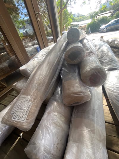 Several rolls of carpets are stacked on top of each other, wrapped in clear plastic. A label is visible on one roll, detailing "Quality, Design, Colour, Size, Carpet" and "HAND MADE IN INDIA." Natural light filters through a window, reflecting off the plastic and the glossy wooden flooring where the carpets are arranged. A parked car and greenery are visible outside the window.