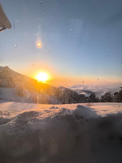 The image displays a winter mountain landscape at the Amirsay ski resort in Uzbekistan, observed through a windowpane at either sunrise or sunset. The sun, a brilliant yellow-orange orb, is positioned low on the horizon to the left of center, partially concealed by a prominent, snow-covered mountain peak. Strong lens flare emanates from the sun. The sky transitions from a clear, light blue overhead to warm orange and yellow hues near the horizon. The foreground consists of substantial, undisturbed snowdrifts, reflecting the warm sunlight. Numerous water droplets and faint streaks are visible on the interior surface of the glass pane through which the image was captured. A light-colored architectural element is partially visible in the upper-left corner. Below the sun-drenched peaks, a thick layer of white clouds forms a vast inversion, resembling a 'sea of clouds,' with the dark, snow-dusted canopies of evergreen trees and distant mountain ridges visible emerging from it. No human figures or active subjects are present in the scene.