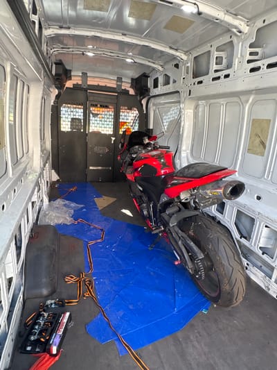 The image depicts the interior of a white cargo van, likely parked in Citrus Heights, California. A red and black sport motorcycle is positioned inside the van, resting on a blue tarp spread across the floor. The tarp is partially covered by clear plastic sheeting and orange and black tie-down straps, suggesting the motorcycle is being prepared for transport. A black case containing tie-down ratchets is visible on the floor to the left of the motorcycle. The van's interior appears utilitarian, with bare metal walls and ceiling, and a caged partition at the rear. Sunlight streams through the rear doors, illuminating the scene.