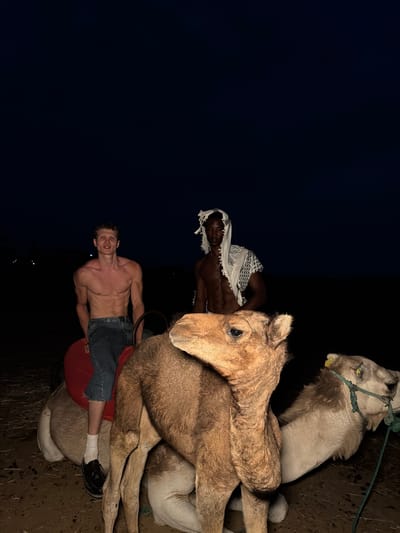 In Douar Ait Daoud, Morocco, at night, two men are seated on camels. The man on the left, shirtless and wearing blue jeans, is facing forward. The man on the right, also shirtless and wearing a white checkered headdress, is looking to his left. In the foreground, the heads and necks of two camels are visible. The camel on the left is closer and its head is turned towards the viewer. The camel on the right is further away and its head is turned to the left. The background is dark, suggesting it is nighttime.