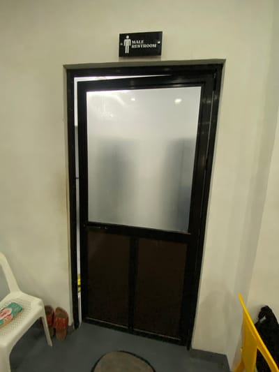 The image depicts a narrow interior hallway or passage, likely in the Philippines given the location context. The primary subject is a closed door with a dark frame and a frosted glass panel on the left half and a dark solid panel on the right half. To the left of the door, a black rectangular sign with white text and a pictogram is mounted on the wall. The text on the sign reads "MALE RESTROOM" and features a stylized human figure.

The wall appears to be painted a light, neutral color, possibly off-white or a very pale yellow. In the upper right corner of the image, a bright yellow object, possibly a plastic chair or a similar item, is partially visible, casting a shadow on the wall. Below the door, on the right side, a white plastic chair with a perforated seat is visible, and on a small shelf to its right, a pair of red and brown sandals are placed. The lighting suggests an indoor setting, likely during daytime, with artificial light contributing to the overall illumination. The overall impression is of a functional, utilitarian space, possibly a public restroom area or a service corridor. There are no people visible in the image, and the scene is static, indicating a moment frozen in time rather than an active event.