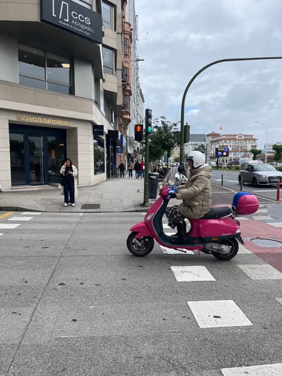 The image captures a street scene in La Coruña, Spain, on an overcast day. A person wearing a white helmet, brown jacket, and checkered pants is seated on a pink moped at a crosswalk, with a pink and blue storage container on the back. The traffic light displays green, indicating it is safe to cross.
In the background, buildings line the street, including one prominently displaying the sign "CCS Abogados Madrid | A Coruña | Vigo" above a doorway labeled "GRAND azulmarino". A woman standing in the doorway appears to be talking on her phone. Other pedestrians are seen further down the sidewalk. A silver SUV is visible across the street with the registration "9044 LNB".