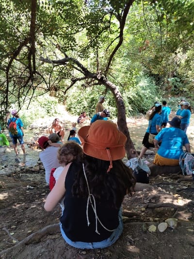The image displays an outdoor scene within a densely wooded area in the Maale Yosef Regional Council, Israel. Approximately 15 to 20 individuals, encompassing both adults and children, are distributed across the frame.
In the foreground, a person with long, dark hair, wearing an orange wide-brimmed hat, a black tank top, and blue jeans, is seated on the dirt ground with their back to the viewer. They are holding a small child with curly brown hair, also facing away. A white, looped cord hangs from the seated person's back. To their right, on the dirt ground, a slight arc of four light-colored, smooth stones is visible, alongside exposed tree roots.
The mid-ground features a shallow stream or river with visible rocks and pebbles on its bed. Multiple individuals are positioned near or in this water body. To the left mid-ground, an adult in a white t-shirt and red cap is seated near the water's edge, appearing to interact with a child. On the right side of the mid-ground, a group of adults, many dressed in light blue t-shirts, are seated on the ground or on natural elements like roots or logs. One adult in a blue t-shirt and a yellow-orange skirt is prominently seated. Scattered bags and personal items are visible near these seated individuals. Further into the background, additional people are seen near the water's edge.
The environment is characterized by lush green foliage and numerous trees, providing extensive shade. A tree with a distinctively curved trunk and exposed root system is prominent in the left mid-ground. The ground consists of uneven dirt, pebbles, and exposed tree roots. The scene is well-lit by natural light filtering through the tree canopy.