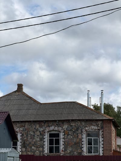 The image shows the upper portion of a rustic, two-story building constructed with stone and brick, located in Byahoml, Belarus. The building has a slate-tiled roof with a brick chimney on the left side and two modern metal chimneys on the right. There are two windows visible on the stone section of the house, both with white frames and multiple panes. In front of the building, a red corrugated metal fence is present. The sky above is overcast with large, white clouds, and three dark power lines are strung across the top of the frame. To the left of the main building, a section of a smaller, grey metal-clad structure with a dark red roof is visible.