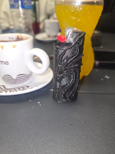 The image captures a close-up, slightly angled view of several objects arranged on a dark, textured surface in Kraishte, Bulgaria. Dominating the center is a black lighter housed in a custom casing with an intricate, embossed design resembling scales or a stylized dragon motif. The lighter features a silver metal top and a red ignition button.
To the left, a clear glass partially holds a vibrant yellow liquid, likely a beverage such as juice. Below and to the right of the lighter, a white ceramic coffee cup with its handle faces the viewer. The cup, sitting on a matching saucer, shows faint brown coffee stains inside and is decorated with distinctive brown, wavy line patterns on its exterior and the saucer. Partially visible text on the cup reads "The P" and "ma," possibly part of a brand name.
The background is dark and out of focus, suggesting an indoor setting under artificial lighting. No individuals or specific activities are depicted; the scene is a still life composition of common items.