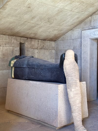 The image depicts an interior scene, likely a film set or exhibit, located in Ouarzazate, Morocco, possibly within Atlas Film Studios. The central focus is a large, dark, sarcophagus-like structure with a textured, matte finish, resting on a sturdy, light grey concrete pedestal. The sarcophagus lid is partially open at the head end, revealing interior details with gold and green accents. To the right of the sarcophagus, a standing mummy figure, wrapped in light-colored, crinkled bandages, is positioned upright, its lower body partially obscured by the sarcophagus and its pedestal.
The surrounding environment consists of walls constructed from light-colored, rectangular stone blocks, mimicking ancient Egyptian architecture. A small, dark, rectangular alcove is cut into the left wall. To the far right, an ornate doorway or archway is visible, adorned with carved hieroglyphs and an Eye of Horus symbol above the lintel. The ceiling is also composed of similar light-colored stone slabs, exhibiting horizontal grout lines. The scene is illuminated by ambient light, casting minimal shadows and suggesting an open-air or well-lit interior setting. No individuals are present in the scene.