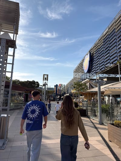 This image, rotated 90 degrees clockwise, captures an outdoor pedestrian area, likely part of a modern entertainment or commercial complex in Paterna, Spain. The setting is bright and sunny under a clear blue sky with scattered wispy clouds.

In the foreground, two individuals walk away from the camera. On the right, a person with long brown hair wears a tan jacket and blue jeans, holding a smartphone in their right hand. To their left, another person, with shorter hair, sports a blue t-shirt with a large white sun-like graphic on the back, light grey sweatpants, and holds a small, bright green object in their left hand. Further in the background, several other people can be seen walking.

The architectural style is modern, featuring buildings with large glass panels, metal frameworks, and structured facades. The ground is paved with a mix of square and rectangular tiles, incorporating a visible drainage grate. To the right of the walkway, a colorful section of what appears to be a carousel or other amusement park ride is partially visible. A prominent circular sign on a building to the left features the words "WORLD" at the top, "PLAY" with an orange burst effect in the middle, and "CENTER" at the bottom, forming "WORLD PLAY CENTER". In the far background, additional structures, including a tall, open-framework tower, suggest the presence of an amusement park or similar attraction.

The bright sunlight casts long shadows, indicating either late morning or late afternoon. On the back of the blue t-shirt, below the sun graphic, the words "STAY" and "RELATED" are clearly visible.