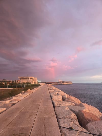 The image shows a paved walkway lined with large rocks on one side and a body of water on the other. In the distance, there are buildings and a faint rainbow in the sky. The sky is filled with pink and purple clouds, suggesting it is either sunrise or sunset. The scene appears to be a coastal area. Based on the architecture and coastal setting, this could be the Lungomare Adriatico in Chioggia, Italy.