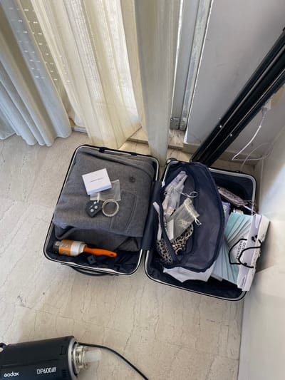 The image displays two open suitcases, suggesting a packing or unpacking activity, set indoors on a light-colored tiled floor next to a window with white blinds or curtains. A white wall forms the background.
The upper suitcase contains a white paper bag with the Greek text "ΟΠΤΙΚΑ ΠΑΝΑΓΟΥΛΙΑΣ" (Panagoulias Optics) prominently displayed, alongside a fan of light blue/white paper or fabric swatches. Inside a navy mesh organizer within this suitcase are smaller pouches, a leopard print item, clear plastic bottles, and other small accessories.
The lower suitcase holds a neatly folded grey garment, likely a jacket or thick shirt. Placed on top of this garment are a small white box with the partially visible text "TO FORM" (likely "TRUE TO FORM"), a silver-colored bracelet, and a black keyring featuring small white stars. An orange-handled lint roller is also visible inside this suitcase.
To the right of the suitcases, a black professional photography strobe light, branded "Godox DP600III," is visible on the floor. A white power cable runs along the wall near black metal tracks, possibly for blinds or a curtain rod. The scene is brightly lit, indicative of daytime.