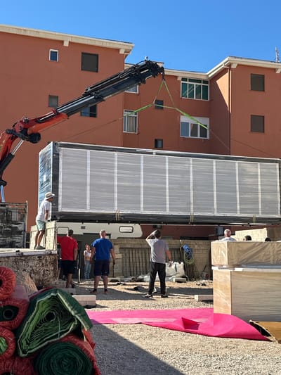 The image shows a scene in the Urbanització Platja De Piles, Spain. A large metal container covered with a white mesh material is being lifted by a crane with an orange and black arm. The container is positioned between a multi-story salmon-colored building with various windows and a truck bed. Several people are present: one wearing a white shirt and straw hat in the truck bed, a man in a blue shirt, a person in a red shirt, and a man in a gray shirt standing on the ground. Rolls of artificial turf in shades of green and red are visible in the foreground, along with a sheet of pink material. Additionally, packaged items are present next to the container.