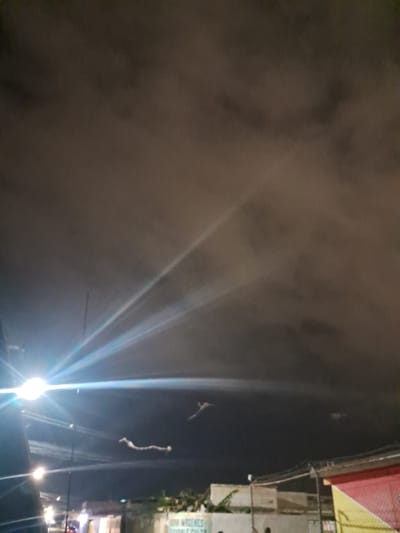 In the city of Cumana, Venezuela, at night, a dramatic sky is illuminated by bright streetlights. The clouds, heavy and dark, are streaked with the intense beams of light, creating a sense of depth and movement. A long, white object, possibly a kite or a piece of fabric, floats across the sky, catching the light. In the distance, urban structures and vegetation are dimly visible, hinting at the city below. The overall scene evokes a sense of wonder and mystery, with the artificial lights clashing with the natural elements of the night sky.