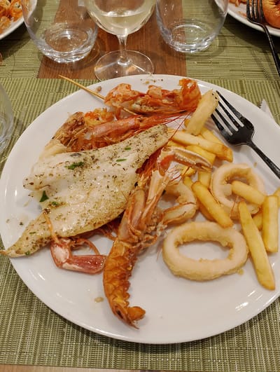 The image shows a white plate of food placed on a green woven placemat on a wooden table. The food consists of fried calamari rings, french fries, and several cooked prawns or langoustines, along with a piece of grilled fish seasoned with herbs. A black fork rests on the plate. In the background, a glass of white wine and an empty glass are visible. The setting appears to be a restaurant in Torri in Sabina, Italy.