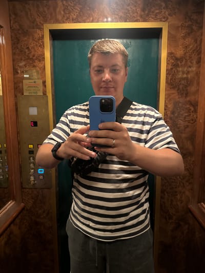 The image shows a person taking a mirror selfie in an elevator. The person is wearing a blue and white striped t-shirt and dark pants. They are holding a blue smartphone to take the picture. The elevator walls have a wood grain pattern and a metallic accent. On the elevator wall, there is an elevator control panel with buttons and a digital display showing the number "3". There is also some text on the elevator wall, but it is not legible. The lighting suggests it is indoors. The overall scene is that of a person using their phone to capture their reflection in an elevator, a common practice for documenting one's presence in a location or outfit.