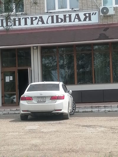 The image shows the rear of a white sedan parked on a gravel lot in front of a building. The car has a license plate that reads "P 310 XK 125". The building appears to be a commercial establishment, with large windows and a sign above the entrance that says "ЦЕНТРАЛЬНАЯ" (Central). An air conditioning unit is mounted on the exterior wall of the building. The scene is likely in the city of Lesozavodsk, Russia, as indicated by the Cyrillic lettering on the sign. The environment is outdoor, with visible trees and foliage in the upper left corner of the image. The car is the primary subject, and the building serves as the background. There are no people visible in the image, and no specific actions or interactions are depicted. The image appears to be a snapshot of an ordinary day in a Russian city.