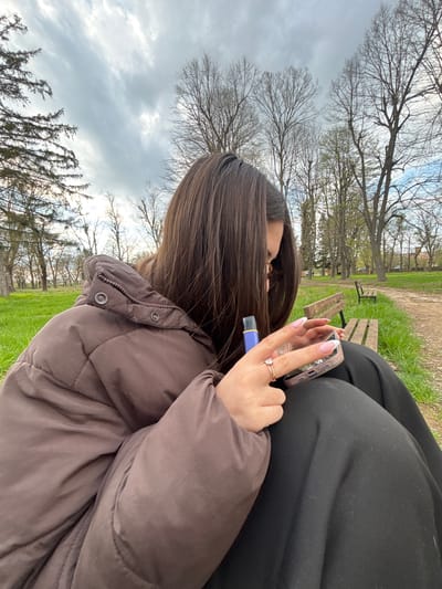 A medium shot shows a person sitting on a bench outdoors in Gromshin, Bulgaria. The person is wearing a brown, quilted puffer jacket with a high collar and several snaps visible. Next to them is a person wearing black pants. The person sitting is holding a smartphone in their right hand, which has a decorative case. In the same hand is a small, purple rectangular object. The person has manicured nails and is wearing a ring on their ring finger. The location is a park, with a grassy area, trees with bare branches, and several benches visible in the background. The sky is overcast. The image appears to have been taken with a slight downward angle.