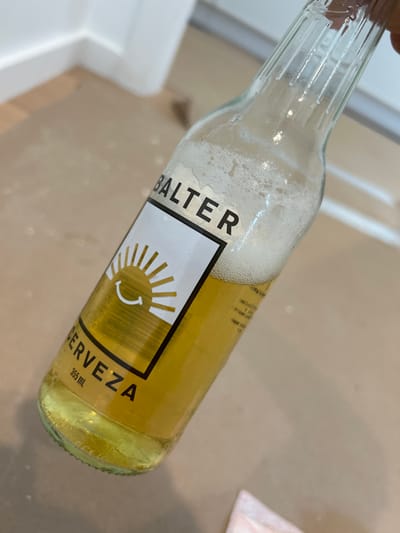 A bottle of Balter Cerveza is tilted diagonally. The clear glass bottle contains a light golden liquid with a foamy head. The label, predominantly white with a black border, features the brand name "BALTER" vertically on the left and "CERVEZA" horizontally on the right, along with the volume "355 mL". A stylized sun graphic with a smiley face is centrally positioned on the label. The bottle is on a light-colored surface.