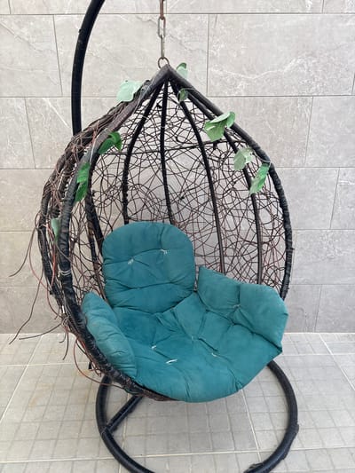 A hanging chair with a teal cushion is situated outdoors, against a wall tiled with grey, marble-patterned squares. The chair is constructed from dark, woven wicker and has a black metal stand. Faux green leaves are entwined around the wicker. The ground is paved with similar grey square tiles. The scene is located in Ayn al Basha, Jordan.