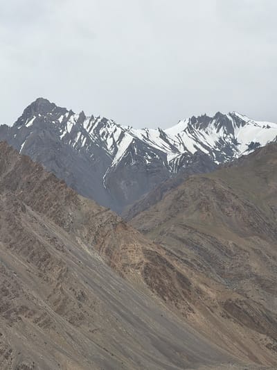A mountainous landscape unfolds under a cloudy sky, with snow-capped peaks dominating the background. The foreground features rugged, layered slopes of rock and earth, displaying a range of earthy tones from brown to gray. The geology suggests a history of significant tectonic activity, evidenced by the folded and faulted strata. The scene is devoid of human presence or any man-made structures, indicating a remote and wild environment. The lighting is diffused due to the overcast conditions, casting a soft, even glow across the terrain and minimizing shadows. The scale of the mountains is immense, conveying a sense of grandeur and isolation. This is a natural scene, captured in the high-altitude region of Zangla, India, a place known for its stark, breathtaking mountain scenery.