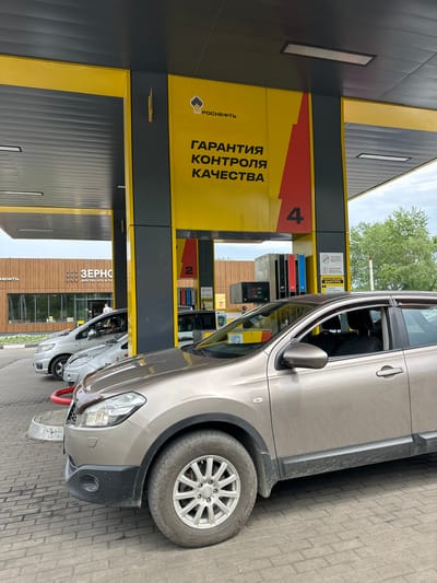 The image shows a gas station with cars lined up to refuel. A prominent yellow and black sign displays the "Rosneft" logo and text in Russian, including "Гарантия контроля качества" (Quality control guarantee) and the number "4". Another sign on the gas pump has the number "2". In the background, a building with "Зерно" (Grain) written on it is visible. A few people are present, including drivers in their cars and a person standing near the building. The scene appears to be a typical gas station environment, with cars, pumps, and associated signage. The location is identified as Slyudyanka, Russia.