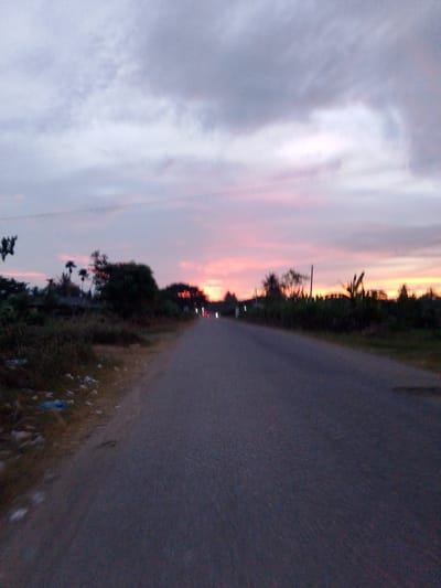 The image captures a dusk scene on a paved road in Glokaron, Indonesia. The sky is overcast with muted shades of gray and lavender, transitioning to hues of pink and orange near the horizon, indicating the setting sun. Silhouetted against the sky are dense foliage and scattered trees along the right side of the road. Several distant, blurred lights are visible on the right side of the road, suggesting vehicles or possibly streetlights. The left side of the road features a dry, grassy embankment littered with debris, including plastic bags. The road itself is dark and appears to be a two-lane street. The overall atmosphere is tranquil and rural.