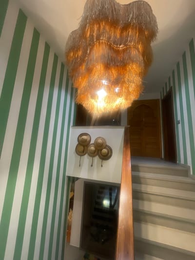 A brightly lit interior space is captured with a slightly rotated perspective. The walls are adorned with horizontal stripes of green and white. A decorative light fixture hangs prominently from the ceiling, emitting a warm, golden glow. On one wall, a collection of straw hats is displayed. A wooden cabinet or door is visible near the top of the image. The location is Tassoultante, Morocco.