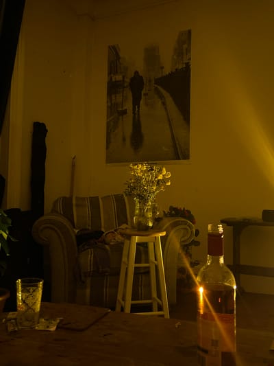 Indoor scene. A black and white photograph of Dundee’s train station hangs on the wall. A sofa is centered in the frame. A bottle lies on a table in the foreground. A glass and other objects are also on the table. A vase of flowers is on the sofa. A chair is behind the sofa.