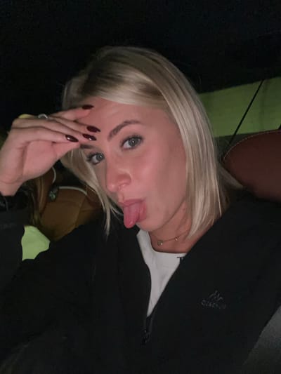 A woman with blonde hair, light skin, and blue eyes is sticking her tongue out. She is wearing a black jacket and a white t-shirt, with a delicate gold necklace. Her nails are painted a dark red. She appears to be indoors and sitting down.
