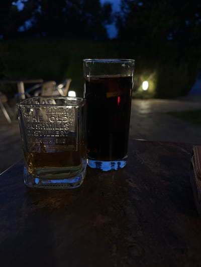 Two glasses of drinks are on a wooden table outdoors at dusk. The glass on the left is a short, square-shaped tumbler containing a golden liquid, possibly whiskey, with some text embossed on its side. The glass on the right is a tall, cylindrical glass filled with a dark carbonated beverage, possibly cola, with a slice of lemon or lime floating within. The background shows dark foliage and out-of-focus lights, suggesting an evening setting in a garden or outdoor seating area. The location is the city of Dragoman, Bulgaria.