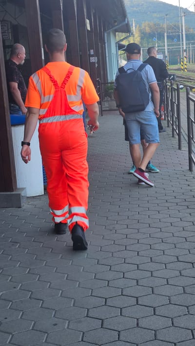 In Kysak, Slovakia, at a train station, a man wearing an orange high-visibility jumpsuit walks away from the camera on a hexagonal tiled platform. He has dark hair and is wearing black boots. He is holding a set of keys in his right hand. To his left, another man in a dark patterned shirt and jeans is partially visible. Further to the right, two men are walking away from the camera. The man closer to the foreground is wearing a light blue polo shirt, light blue shorts, and a dark backpack, with red and white sneakers. The man behind him is wearing a dark jacket and dark pants. The background shows a train track, a station building, and trees on a hillside.