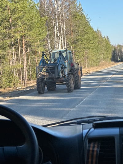 The image captures a rural outdoor scene, likely along a road in a forested area, with the location context provided as Krasnoyar, Russia.
The primary subject is a heavy-duty, blue or dark green forestry machine, resembling a forwarder or skidder, positioned on the shoulder of a paved road. It features large, deeply treaded tires indicative of off-road capability and rugged construction suitable for logging operations. The vehicle has a prominent hydraulic arm structure at its rear, although no logs are currently being carried. Visible text on the back of the machine appears to be "BROUTTE" and possibly "1010" or "1010T," suggesting it might be a Bruunett forestry vehicle.
The road is asphalt, with long, distinct shadows cast across it, hinting at either morning or late afternoon. The sky is clear and blue, indicating bright, sunny weather. To the left of the road, a dense forest stretches into the background. This forest comprises both coniferous trees, primarily pines with green foliage, and deciduous trees, likely birches, with their characteristic white bark and bare branches. Many felled logs are visible among the trees, reinforcing the forestry context of the scene.
The photograph is taken from inside another vehicle, with a portion of the dashboard and a steering wheel visible in the lower right foreground. Notably, a St. George ribbon, a symbol of military valor and patriotism in Russia, is affixed to the dashboard of the viewer's vehicle, providing a cultural detail. The activity appears to be the viewer driving past or stopped near the forestry machine, which itself might be parked or moving slowly.