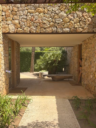 The image shows an outdoor entryway at a stone-walled residence in Artà, Spain. The entryway is a covered walkway leading from a sunlit outdoor area into a shaded interior. The walls are constructed of rough-hewn, natural stones in various earthy tones. In the foreground, a pathway of light-colored gravel leads towards the entrance. To the left, a young woman in a black bikini top and blue bottoms stands in a doorway, leaning against the stone wall. She appears to be looking out into the outdoor space. The outdoor area features lush green foliage, including palm trees and a neatly trimmed hedge. A rustic wooden bench is visible in the middle ground. The lighting suggests it is daytime, with strong sunlight casting shadows.