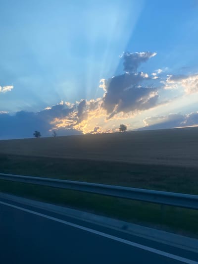 The image shows a wide shot of a rural landscape under a dramatic sky. The sun is setting, with bright rays of light piercing through dark, textured clouds. The clouds are illuminated from behind, creating a golden glow in some areas. Below the sky is a gentle, rolling hill covered in dry, golden-brown grass, suggesting a field of crops or hay. Two solitary trees are visible on the crest of the hill, silhouetted against the bright sky. In the foreground, a portion of a road is visible, with a metallic guardrail running horizontally across the bottom third of the image. The road appears to be paved with dark asphalt and has a white line marking its edge. The overall impression is one of a serene, possibly late summer or autumn evening in a rural area. The scene is devoid of people or any discernible activity, focusing solely on the natural elements of the landscape and the atmospheric lighting. There is no specific indicator of the city of Svetlîi, Moldova, in the image.