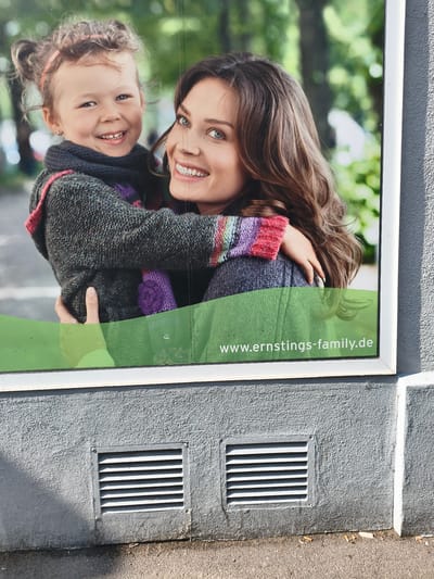 This media file shows a close-up of a poster featuring a woman and a young child. The woman, with long, wavy brown hair and bright blue eyes, is smiling warmly. She is wearing a grey knitted sweater with colorful striped cuffs and a grey scarf. The child, also smiling, is nestled into the woman's side, wearing a matching grey knitted sweater and a purple knitted mitten. The background of the poster is blurred greenery, suggesting an outdoor setting, possibly a park.

Below the image of the woman and child, there is a green wave-like graphic. Visible on this green area is white text that reads "www.ernstings-family.de". The poster appears to be displayed on the exterior of a building, as indicated by the textured grey wall below it, which features two louvered air vents. The lighting suggests it is daytime, with sunlight casting shadows on the wall. The presence of cigarette butts on the ground near the wall implies this is an outdoor public space. The overall emotion conveyed is one of warmth, family, and happiness. The location context is Lörrach, Germany.