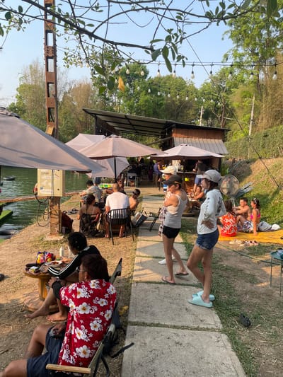 This image captures a lakeside scene in Tambon Nam Phrae, Thailand. Several people are engaged in different activities. Some individuals are seated under umbrellas, appearing to be dining or relaxing. Others are sunbathing on the grass, while a couple is walking away from the main group. An outdoor cafe or restaurant is present, with tables and chairs set up for patrons. A body of water is visible, with some form of electric equipment located near the shoreline. The environment includes lush greenery and trees, indicating a natural setting.