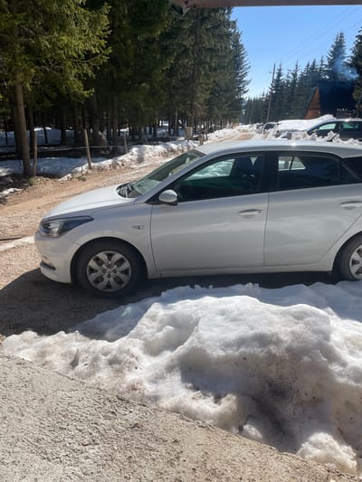 A white four-door hatchback vehicle, consistent with a Hyundai i20 model, is parked on the dirt and gravel shoulder of a snow-lined road in Palež, Montenegro. The driver's side front door of the vehicle is slightly ajar. A significant accumulation of dirty, melting snow rests on the ground immediately adjacent to the car and to the right of the foreground. The road, partially obscured by snowdrifts, extends into the distance through a dense coniferous forest. Patches of snow are visible on the forest floor. In the background on the right, a dark-roofed building, possibly a cabin or lodge, is visible, emitting smoke or steam from its chimney. Overhead utility lines are also present. The sky is clear and blue, indicating bright daylight. No individuals are visible in the scene.