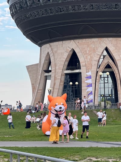 This image depicts an outdoor scene near a large, architecturally distinctive building, possibly a cultural center or museum, characterized by its pale stone facade and arched openings. In the foreground, a massive, inflatable orange dog, resembling a Corgi, is a prominent attraction. A group of people, varying in age and attire, are scattered across a grassy area. Some are standing and conversing, while others are seated or walking. A small dog is being walked on a leash near the inflatable. The background shows more people, some sitting on a ledge overlooking a city skyline. The sky is partly cloudy. The scene suggests a public gathering or event, possibly in a park or plaza in the city of Novyi Poselok, Russia.