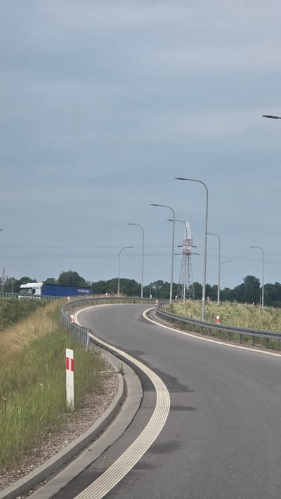 This is a media file showing a view from a road in Suwałki, Poland. The scene is dominated by a curving asphalt road with white lane markings. To the left of the road, there are several tall lamp posts lined up. In the background, a large lattice-like electrical pylon is visible, suggesting the presence of power lines that stretch across the frame. Beyond the lamp posts and pylon, a dense line of green trees forms a natural backdrop. A blue semi-trailer truck with white cab is parked on the side of the road. The weather appears overcast, with a pale, diffused sky. There are some small red and white markers along the side of the road. The image is oriented vertically, as if the camera was turned on its side.