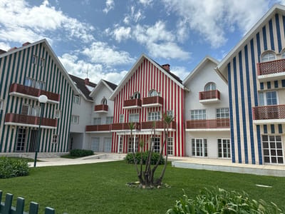 The image displays a cluster of distinctive, multi-story residential buildings located in Ílhavo, Portugal, characterized by their vertically striped facades, reminiscent of the traditional "palheiros" of Costa Nova. Three primary buildings are prominent: one on the left with green and white stripes, a central one with red and white stripes, and another on the right with blue and white stripes. A plainer, white-painted building is recessed in the background between the green and red striped structures. All buildings feature pointed gable roofs, numerous rectangular windows, and ground-level glass doors. Multiple balconies with reddish-brown railings are visible on each building, some surmounted by arched windows. In the foreground, a well-maintained green lawn occupies the central space, bordered by a low dark green fence along the bottom edge of the frame. A single white spherical streetlight on a dark pole stands on the left side of the lawn. Patches of green foliage and a multi-stemmed shrub are planted in garden beds within the lawn. The scene is observed under a blue sky with scattered white clouds. No individuals are present.