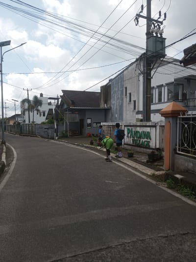 The image depicts a street scene in Ban Phouviengnoy, Laos. The road is paved and curves to the left. On the right side of the road, there is a sign that reads "PANDAWA VILLAGE" in green lettering. Three young people are standing near the sign. One is bending over, and another is wearing a blue shirt and a backpack. Buildings are visible in the background, including a two-story white building with gray trim and a gray-roofed structure. Overhead, there are numerous power lines and utility poles. The sky is overcast with clouds. Vegetation is growing along the roadside. A street light is visible on the left side of the road.