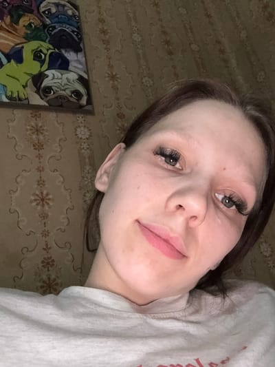 A woman with brown hair and false eyelashes is taking a selfie. She is wearing a light-colored sweatshirt. Behind her, a patterned wallpaper is visible. To the left of the woman, there is a painting of pugs. The image appears to have been taken indoors.