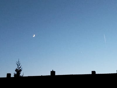 This image shows a clear blue sky with a crescent moon visible in the upper left quadrant. A small, bright celestial object, possibly Venus, is visible in the upper right quadrant. Silhouetted against the sky are the rooftops of buildings, with a chimney prominently featured on the left and another smaller chimney in the center. A solitary tree is also silhouetted to the left of the center chimney. The scene appears to be captured at dusk or dawn, given the lighting conditions and the visible moon. The location is Kitzingen, Germany, as confirmed by the presence of characteristic German rooftop architecture.