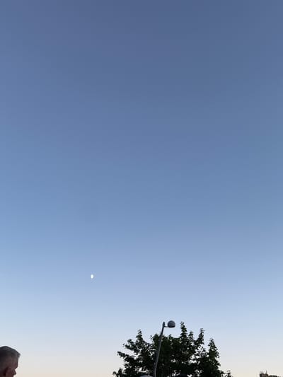 This is a vertical, medium shot image of a clear blue sky with a crescent moon visible in the upper left quadrant. In the bottom of the frame, the top of a person's head and shoulder are visible on the left. To the right of the person, a streetlight stands in front of a cluster of green trees. The image appears to have been taken in Jonava, Lithuania, likely during the evening as indicated by the moon's visibility and the transition from daylight to dusk in the sky gradient.