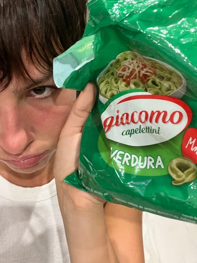 A person is holding a green package of "Giacomo capelettini VERDURA" pasta close to their face. The pasta is vegetable-filled, as indicated by the label. The person has brown hair and is wearing a white shirt.