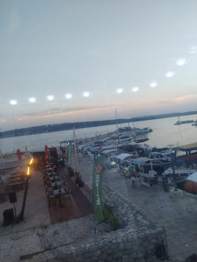 The image depicts a waterfront scene in Crikvenica, Croatia, likely during the evening or twilight hours. A body of water, presumably the Adriatic Sea, is visible in the background, with a distant coastline featuring hills. Along the waterfront, a harbor is filled with numerous boats of various sizes, ranging from small motorboats to larger vessels.
The foreground and midground show an outdoor seating area with many tables and chairs, suggesting a restaurant or cafe. A large green banner with the Heineken logo is prominently displayed. Various structures, possibly kiosks or food stands, are present, along with several people walking around or seated. Some of the tables appear to be set for dining.
The sky is overcast with a pale, diffused light, and there are reflections on the surface from overhead lights, indicating the presence of artificial illumination. The ground is paved with stone or concrete, with some gravel areas and low vegetation. The overall atmosphere suggests a relaxed evening setting in a popular coastal town.