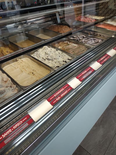 A slightly elevated eye-level, canted perspective captures a commercial freezer display case, laden with various flavors of ice cream. Metal containers hold the ice cream, and each flavor is identified by a label on a red strip along the upper edge of the case. These labels, written in German, include "SAHNE-KIRSCH", "Kinder Country", "SCHOKOKEKS", "RUM-KROKANT", "PISTAZIE", "Stracciatella", "HASELNUSS", and "MOZART". The background is of a contemporary aesthetic, with a light blue panel abutting a wall with large gray square tiles. The overall appearance is modern and clean, indicative of a commercial food establishment. This image was taken in the city of Lünen, Germany.