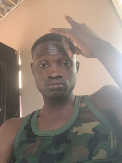 A dark-complexioned adult male is depicted in an indoor setting, posing in what appears to be Kado, Nigeria, though the specific location within the city is not visually discernible. The male, with short, dark hair and a fade haircut, looks directly into the camera with a neutral expression. His right hand is raised, fingers splayed, resting above his forehead. He is wearing a green camouflage-patterned tank top. The background features a plain, light-colored ceiling, with a decorative patterned tile section visible in the upper left corner. To the subject's left, a dark vertical door frame or wall edge is visible, illuminated by a bright vertical strip of light, suggesting an adjacent open space.